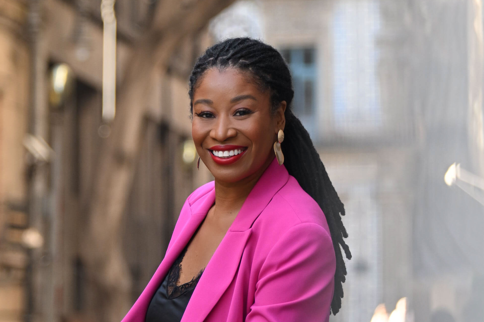 Latifah Abdur smiling in a bright pink blazer against a brick wall