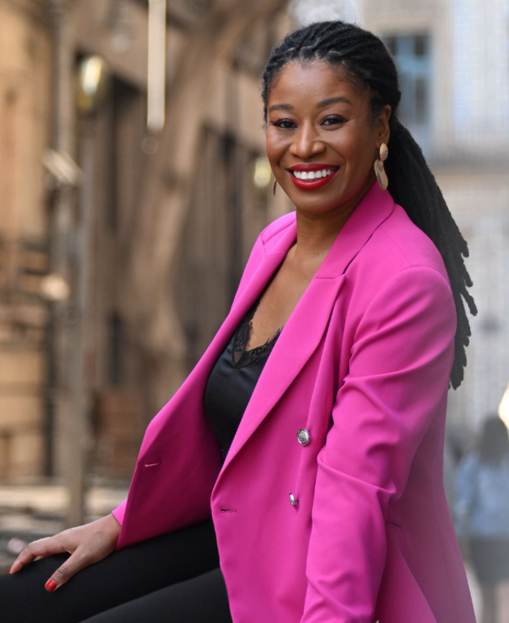 Latifah Abdur smiling in a bright pink blazer against a brick wall